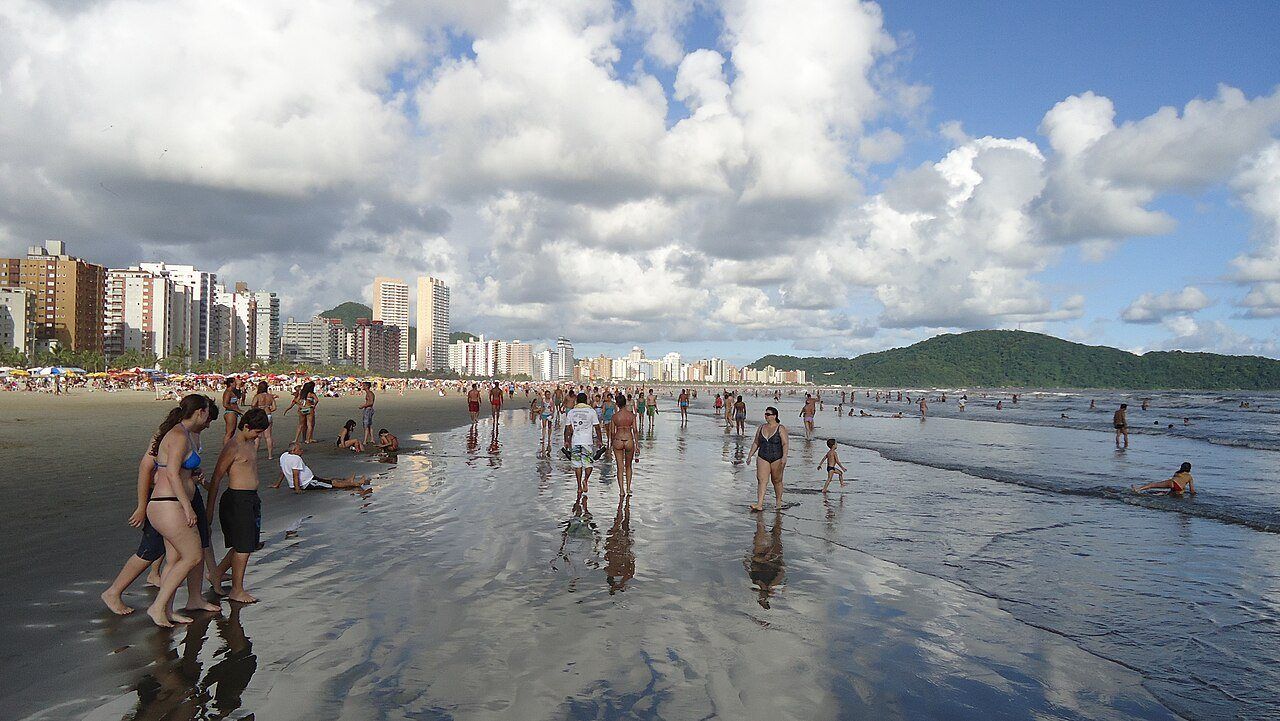 Praia da Guilhermina cheia, com prédios e morro ao fundo