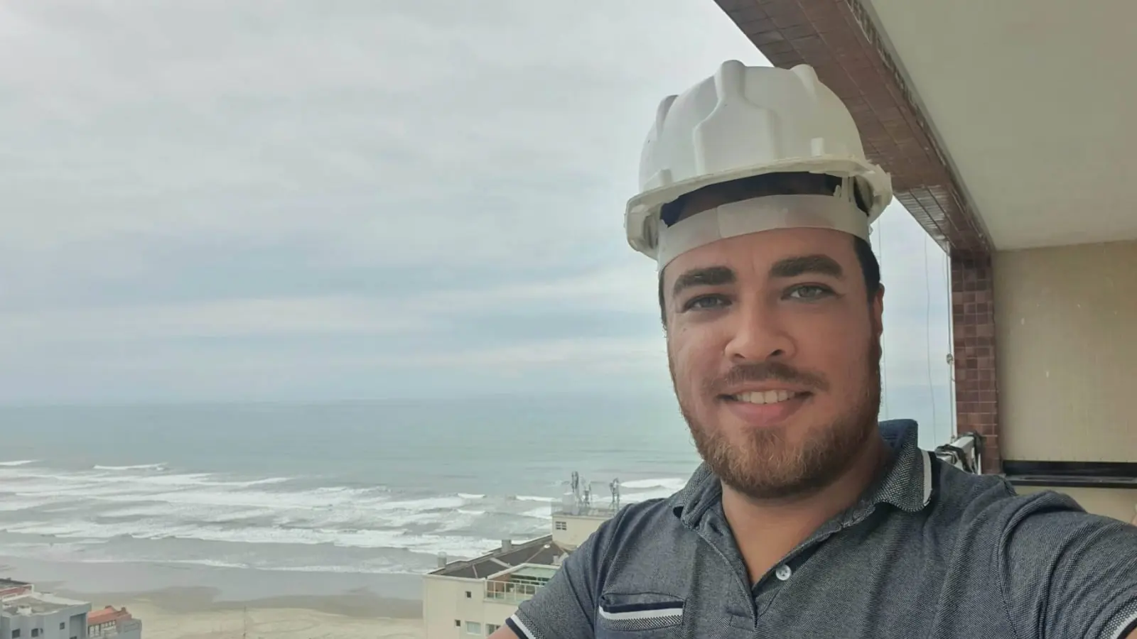 Samuel Laupp, corretor de imóveis, de capacete na sacada de uma obra com o mar de Praia Grande ao fundo